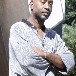 A man standing outdoors with arms crossed, wearing a patterned tunic and necklace, exuding confidence, set against a background of abstract art and greenery.