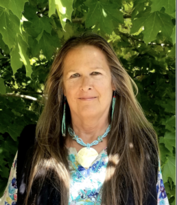 Portrait of a woman with long brown hair, wearing turquoise jewelry and a colorful top, standing in front of lush green foliage.