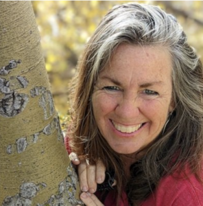 A smiling woman with long, wavy hair partially gray, leaning against a tree trunk in an outdoor setting with golden foliage in the background. She wears a red garment, and her expression radiates warmth and joy.
