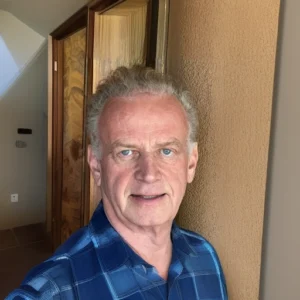 Portrait of James Butcher in a blue checkered shirt standing indoors near a wooden doorway, with natural light highlighting his features