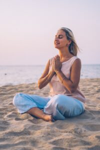 Woman meditating on a sandy beach at sunrise, hands in prayer pose, embracing mindfulness and peace in a serene coastal setting.