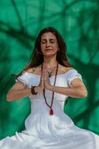 Serene woman meditating with hands in prayer position, wearing a white dress and mala beads, set against a vibrant green background with natural shadow patterns.