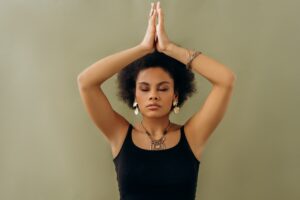 Close-up of a person with natural curly hair holding their hands in a prayer position above their head, wearing bracelets, set against a neutral background