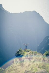 Person standing triumphantly on a mountaintop, arms raised in celebration, with a breathtaking view of misty cliffs and a faint rainbow in the foreground.