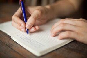 Close-up of hands writing in a notebook with a blue pen, symbolizing creativity, journaling, or note-taking.