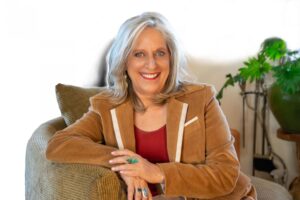 Smiling woman with silver hair sitting on a comfortable chair, wearing a tan blazer and red top, adorned with turquoise and silver jewelry, with green plants in the background.
