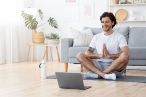 Smiling young man sitting in lotus pose in living room in front of laptop, meditating. Sporty guy having yoga class online, sitting on yoga mat and looking at laptop screen in living room