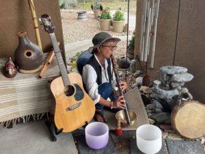 Glee Briggs engaging in a musical meditation session outdoors, playing a flute surrounded by an acoustic guitar, singing bowls, chimes, and other musical instruments, with a small water fountain in the background.