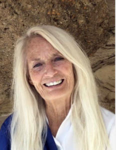 Close-up of a smiling woman with long blonde hair, standing against a natural rock background, radiating warmth and joy.