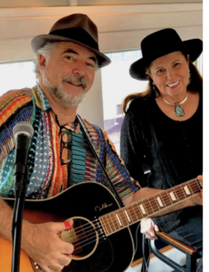 Marc and Paula Rhae performing together, with Marc playing the guitar and Paula smiling beside him, both wearing stylish hats and exuding a warm and creative energy.