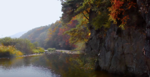 Scenic autumn landscape featuring vibrant fall foliage, a calm river reflecting the colorful trees, and rocky cliffs under a clear sky, creating a serene and picturesque view.