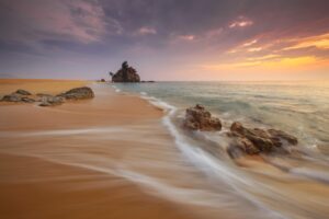 Golden sandy beach at sunset with gentle waves flowing over rocks, vibrant colors in the sky, and a small island in the distance, evoking a sense of tranquility and natural beauty.