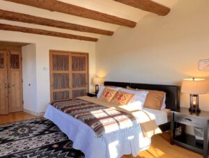 Premium Pueblo Southwestern-style bedroom featuring wooden ceiling beams, a cozy bed with patterned blankets and pillows, rustic wooden doors, and warm ambient lighting.