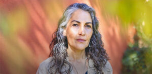 Portrait of Marcela Lobos with silver-streaked hair, standing against a warm-toned backdrop with soft sunlight and natural greenery, evoking a sense of wisdom and calmness.