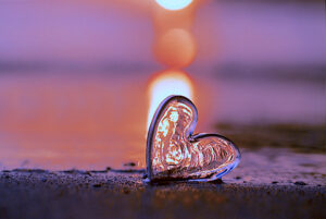 Glass heart on a sandy surface reflecting warm light at sunset, with a glowing background creating a peaceful and romantic atmosphere.