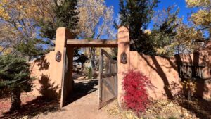 Rustic wooden gate set in an adobe wall surrounded by autumn trees, vibrant red foliage, and clear blue skies, welcoming visitors into a serene Southwest garden.