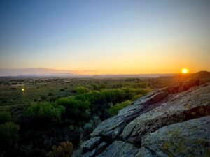 Stunning sunset over a vast Southwestern landscape with rolling hills, lush green trees, and rocky foreground, casting a golden glow across the horizon.