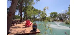 Serene lakeside deck with red Adirondack chairs, shaded by tall trees, overlooking a peaceful pond with a water fountain, surrounded by lush greenery under a clear blue sky.