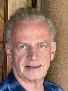 Close-up portrait of James Butcher, wearing a blue checkered shirt, standing near a wooden door with a neutral background.