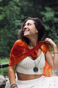 Julie Shapiro smiling joyfully outdoors, wearing a vibrant orange scarf, a white crocheted top, and a green stone pendant necklace, surrounded by lush greenery in a serene natural setting.
