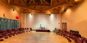 Interior of the Kiva, a spacious circular meeting room with wooden beams, arranged chairs, meditation cushions, and a serene altar, creating a peaceful and welcoming space for gatherings and practices.