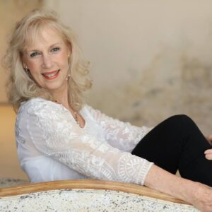 Portrait of Lynda Hope sitting in a relaxed pose, smiling warmly, wearing a white embroidered blouse and black pants against a neutral background.