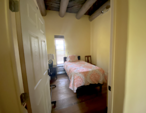 Cozy bedroom in Main Lodge #6 - MOTHER EARTH, featuring a single bed with a peach-patterned comforter, a bedside table with a decorative lamp, and natural light streaming through the window, highlighting the rustic wood-beamed ceiling.
