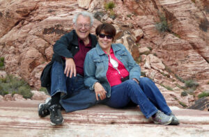Happy couple sitting together on rocky terrain, surrounded by striking red rock formations, enjoying a moment of laughter and connection in nature.