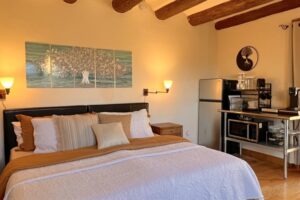 Interior of Pueblo #2 CHACO featuring a cozy bedroom with a king-sized bed, wooden ceiling beams, kitchenette with appliances, and wall art depicting a serene tree design.