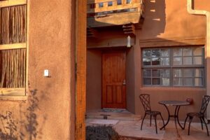 Exterior view of Pueblo #3 featuring a wooden front door, a cozy patio with wrought iron table and chairs, adobe-style walls, and warm sunlight casting soft shadows