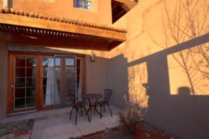 Exterior view of Pueblo #4 featuring a private patio with a small wrought iron table and chairs, adobe-style walls, wooden pergola, and glass doors reflecting the warm sunlight.