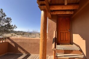 Exterior view of Pueblo #8 featuring a wooden front door, rustic wooden beams and columns, a sunlit patio, and scenic desert landscape in the background.