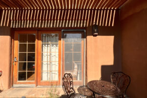 Patio view of a serene adobe-style building with wooden French doors, a glass screen door, and a wrought iron table and chairs under a pergola casting shadow patterns.