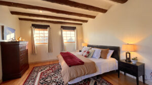 Cozy, Southwestern-inspired bedroom featuring exposed wooden beams, a large bed with warm-toned bedding, and a vibrant area rug. Natural light flows through two framed windows, complementing the room's serene, rustic charm. A sleek black nightstand with a lamp adds a modern touch alongside a classic dark wood dresser.