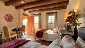 Pueblo House #8 - TAOS: Bright bedroom with exposed wooden beams, two cozy beds adorned with red accents, a sitting area with chairs and table, fresh flowers, and a welcoming open door leading to natural light.