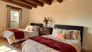 Bright and cozy bedroom featuring two beds with plush, patterned bedding, red throws, and decorative pillows. Natural light streams through the windows, complementing the rustic wooden beams and soft neutral tones of the space. A vase of vibrant flowers decorates the dresser, adding warmth to the room's serene ambiance.