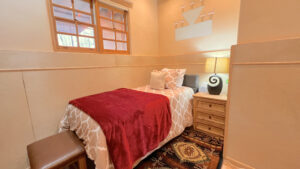 Serenity Center Room #5 - PURITY at the Southwest Sanctuary. Cozy single room with a warm red throw blanket, soft beige bedding, natural wood nightstand, and a decorative wall mural. Relaxing and peaceful retreat space for rejuvenation.