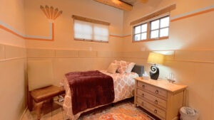 Cozy bedroom at The Serenity Center featuring a single bed with plush red and beige bedding, decorative pillows, and warm lighting. A wooden nightstand and chair complete the peaceful atmosphere for rest and relaxation.