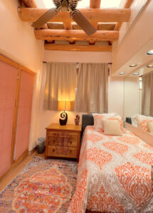 Serenity Center Room #1 HARMONY with single bed, bright orange and white bedding, wooden nightstand, decorative lamp, and built-in mirror wall. Features a maroon sink and cozy Southwestern design with warm wooden ceiling beams and neutral curtains.