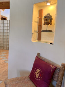 Cozy corner in the Serenity Center featuring a rustic chair adorned with a decorative red pillow, a niche display with a golden Buddha head sculpture, and a softly lit ambiance that evokes tranquility and mindfulness.