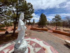 Statue of Guan Yin in a serene outdoor setting surrounded by pine trees and a red earth pathway under a partly cloudy blue sky at the Southwest Sanctuary.