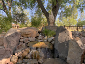 Serene outdoor setting featuring a statue of a spiritual figure hanging near a large tree, surrounded by lush greenery and dappled sunlight against a clear blue sky.