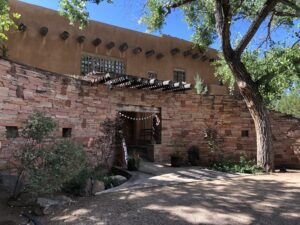 Main Lodge Entrance featuring adobe-style architecture, rustic stone walls, wooden pergola accents, and lush greenery under a large, shaded tree against a clear blue sky.
