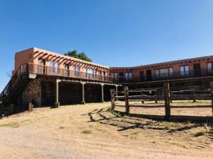 Exterior view of The Lofts & Stable Rooms featuring adobe-style architecture, wooden balconies, rustic fencing, and a clear blue sky, showcasing a Southwestern retreat setting.