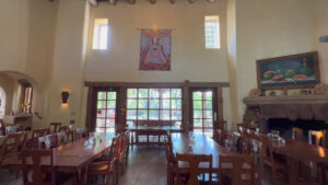 Spacious dining hall with wooden tables and chairs, large windows, a stone fireplace, and decorative artwork on the walls, creating a warm and inviting atmosphere.