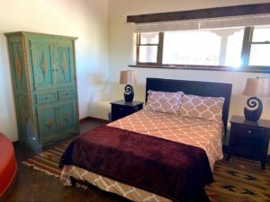 RanchHouse #3 - BANCOS room at The Chi Center showcasing a cozy bed with geometric bedding, a rustic green armoire, traditional southwestern decor, and natural light streaming through large windows.