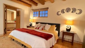 Elegant Pueblo House #9 - TESUQUE room at The Chi Center, featuring a king-size bed with luxurious linens, handcrafted wood accents, moon phase wall art, warm ambient lighting, and a private en-suite bathroom for a serene and restorative retreat.
