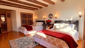 Cozy Pueblo House #3 - COCHITI room at The Chi Center, featuring two comfortable beds with warm bedding, wooden ceiling beams, natural light, and elegant rustic decor for a peaceful and inviting retreat experience.