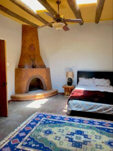 RanchHouse #2 - PLACITAS room at The Chi Center showcasing a traditional adobe-style fireplace, rustic wooden ceiling beams, a vibrant southwestern area rug, and a cozy bed with patterned bedding for an authentic retreat experience.