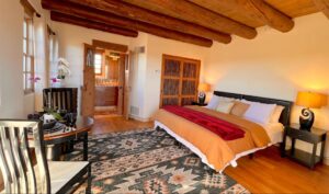 Inviting Pueblo House #6 - NAVAJO room at The Chi Center, featuring a king-size bed with elegant linens, a cozy seating area, tribal-patterned rug, wooden ceiling beams, warm lighting, and an en-suite bathroom for a comfortable retreat.